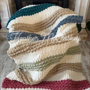 Puede incluir: Una manta de punto grueso con rayas en verde azulado, blanco, beige, gris, verde oliva y burdeos. La manta es suave y acogedora, ideal para el sofá. El texto "Heather McClaskey" está en cursiva.