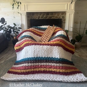 May include: A chunky knit blanket in a variety of colors, including white, teal, burgundy, and gold, is draped over a chair. A wooden knitting loom rests on top. The blanket has a striped pattern and a thick, textured appearance.