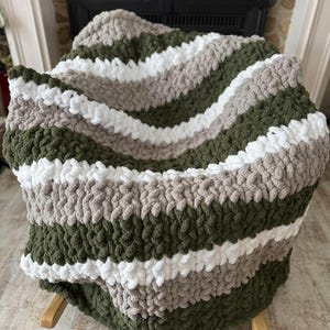 Puede incluir: Una manta de punto grueso con rayas horizontales gruesas en colores alternos verde oliva, beige suave y blanco brillante. La manta está confeccionada con un hilo suave y afelpado con una textura de bucles grandes visible en toda la pieza.