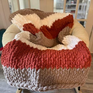 May include: A chunky knit throw blanket featuring horizontal stripes in alternating colours of burnt orange, cream, and light brown. The blanket is crafted from thick, soft yarn with a visible looped texture, draped over a rounded chair frame.