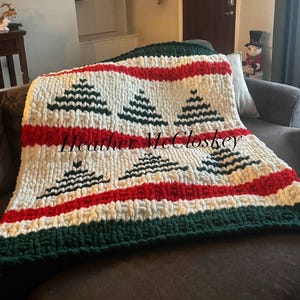 May include: A chunky knit blanket in cream, red, and green, with a repeating pattern of black Christmas trees. The blanket is draped over a sofa. The text "Heather McCloskey" is visible on the blanket.