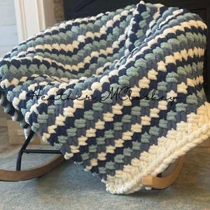 May include: A handmade, chunky knit blanket in a rocking chair. The blanket features a checkerboard pattern in shades of navy, light blue, and cream. The blanket is draped over a wooden rocking chair with a black frame. The text "Heather McCloskey" is visible.