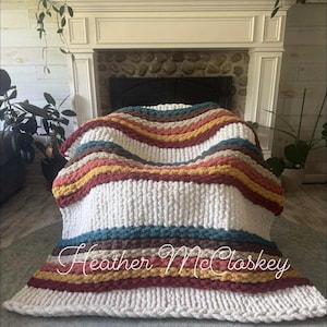 Puede incluir: Una manta de punto grueso con rayas horizontales en crema, turquesa, marrón, amarillo y burdeos. La manta está sobre una silla. El nombre "Heather McCloskey" está escrito en cursiva en la parte inferior.
