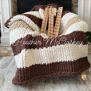 May include: A chunky knit blanket in brown, white, and tan stripes. The blanket is folded over a chair and has three wooden knitting tools resting on top. The blanket has the text "Heather McCloskey" in a cursive font.