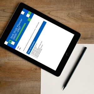 Op de afbeelding: Een zwarte tablet toont het document "NHS Shift Planner 2026-2027", met een blauw en groen kopje. Een zwarte potlood en een blanco wit papier liggen op een houten oppervlak. Het tabletscherm toont een inhoudsopgave.