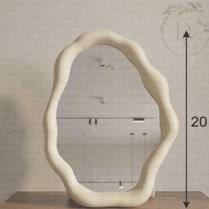 以下が含まれることがあります： 不規則で波打つような有機的な形状のフレームを持つクリーム色の鏡。鏡の横にある垂直の矢印グラフィックは、高さが20cmであることを示しています。鏡面には天井の照明を含む周囲の環境が映り込んでいます。