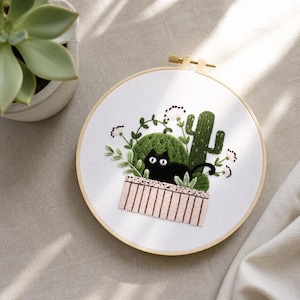 May include: Embroidered hoop art featuring a black cat nestled among green cacti and foliage. The design is set in a white fabric, framed by a wooden hoop. The cat has large, round eyes, and the embroidery includes details like small white flowers.