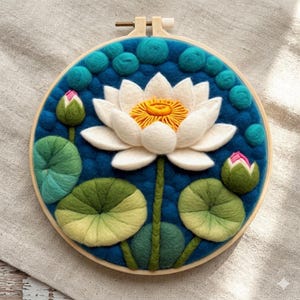 Puede incluir: Una pieza de arte de fieltro hecha a mano con una flor de loto blanca con un centro amarillo, rodeada de hojas y capullos de nenúfar verdes, sobre un fondo azul texturizado dentro de un aro de bordado de madera.
