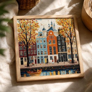 May include: A square framed cross-stitch embroidery depicting a canal-side street scene with colorful narrow buildings, autumn trees with yellow and orange leaves, and a small boat on the water. Several small figures are walking along the sidewalk.