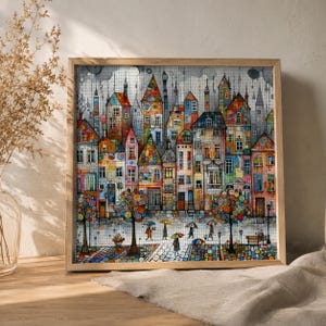 May include: A square framed wall art piece featuring a colorful, detailed illustration of a city street with tall, narrow buildings in various shades of orange, blue, and yellow. The scene depicts people walking with umbrellas under a rainy sky.