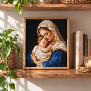 May include: A framed cross-stitch embroidery depicting a woman with a white head covering holding a baby wrapped in a white cloth. The woman wears a blue garment against a black background. The artwork is encased in a light-coloured wooden frame.