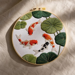 May include: Embroidered artwork featuring four koi fish in a pond with green lily pads. The fish are orange, white, and black, set against a white background within a wooden embroidery hoop.