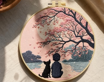 May include: Embroidered hoop art featuring a serene scene of a person and a dog under a cherry blossom tree. The artwork uses pink, blue, and black thread on a light pink background. The design is set in a wooden hoop.