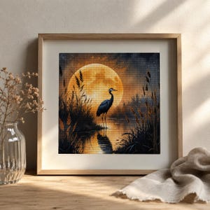 Könnte beinhalten: Ein quadratisches gerahmtes Wandbild mit einem Diamant-Painting eines dunklen Reihers, der nachts in einem Sumpf steht. Ein großer, leuchtender goldener Vollmond erhellt die Szene, wirft Reflexionen auf das Wasser und ist von hohen Schilfgräsern umgeben.