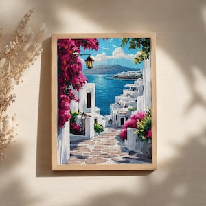 Può includere: Opera d'arte a punto croce incorniciata raffigurante una scena di un villaggio costiero. L'opera presenta edifici bianchi, un sentiero acciottolato e vibranti fiori rosa. Il mare e il cielo blu completano la vista pittoresca. La cornice è in legno chiaro.