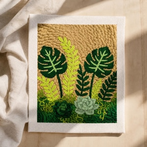 May include: A framed textile art piece with a botanical design. It features green tropical leaves and succulents against a textured tan background. The piece is framed with a white canvas border, creating a vibrant, nature-inspired aesthetic.