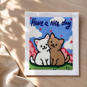 May include: A square, handmade punch needle artwork showing two cartoon cats embracing. The blue background has white clouds and the text "Have a nice day". The cats are white and brown, sitting on green grass.