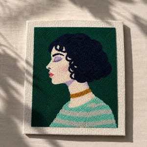 May include: Embroidered artwork depicting a woman's profile. The portrait shows a woman with dark curly hair, wearing a striped green and grey shirt and a gold choker. The background is a deep green, and the piece is framed in a neutral-toned fabric.