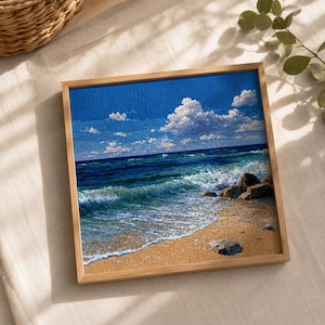 May include: A square cross-stitch embroidery depicting a coastal scene with a sandy beach, turquoise ocean waves crashing against dark rocks, and a bright blue sky filled with white clouds, all encased in a light-coloured wooden frame.