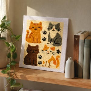 May include: A square, framed artwork featuring four cartoon cats in shades of orange, brown, and gray, with black paw prints. The cats are set against a light yellow background. The artwork is displayed on a wooden shelf with books and a small vase.