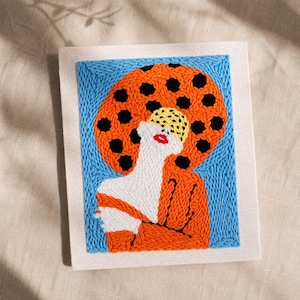 May include: A colorful embroidered artwork featuring a woman with a large orange sun-like shape behind her. The woman's figure is white, with red lips and a yellow and black patterned hat. The background is blue, and the artwork is on a white canvas.