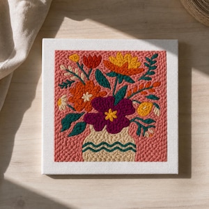 May include: A square, framed floral embroidery. The artwork features a vase filled with colorful flowers in shades of orange, yellow, and purple, set against a textured pink background. The vase is beige with a wavy green design.