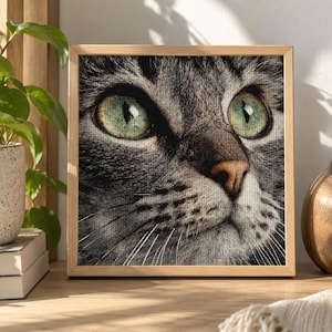May include: A square framed wall art piece featuring a detailed close-up portrait of a grey tabby cat with striking green eyes and a pink nose. The image has a visible cross-stitch texture throughout, set within a light-toned wooden frame.