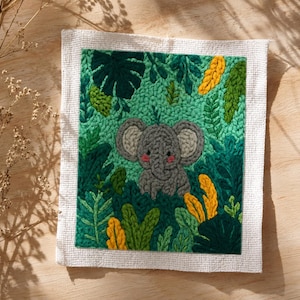 Peut inclure: Broderie textile représentant un éléphant gris aux joues roses entouré d'un feuillage vert luxuriant et d'accents jaunes. L'œuvre est encadrée d'une bordure blanche, créant une scène de jungle vibrante.