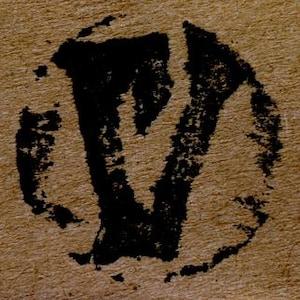 May include: A black ink stamp impression on a textured tan paper surface. The stamp features a bold, stylized letter V enclosed within a circular border. The ink application appears uneven and distressed, showing the natural fibers of the paper.