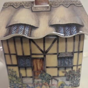Könnte beinhalten: Eine keramische Teekanne in Form eines Cottage, mit einem Strohdach und einem Schornstein. Das Cottage hat gelbe Wände mit braunen Fachwerkkonstruktionen, eine Holztür und kleine Fenster. Der Boden der Teekanne ist mit floralen Mustern verziert.