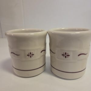 Puede incluir: Dos tazas de cerámica color hueso con forma cilíndrica lisa y borde acampanado. Cada taza está decorada con una línea horizontal de color granate oscuro cerca de la base y una banda central con un pequeño motivo floral de cuatro pétalos.