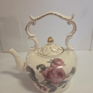 May include: A cream-coloured ceramic teapot featuring a floral pattern of pink roses and green leaves. The teapot has a gold-trimmed handle, a matching lid with a gold-accented finial, and a curved spout. The design includes delicate gold detailing.