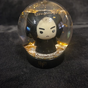 May include: A clear glass snow globe containing a small figurine of a character with black hair and a black robe. The globe is filled with liquid and gold glitter particles. The base is black and features the gold letters HP with a lightning bolt.