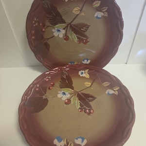 Puede incluir: Dos platos redondos de cerámica con borde festoneado, con un diseño degradado que va desde un borde granate oscuro hasta un centro color canela. Los platos están decorados con ilustraciones pintadas de hojas marrones, bayas rojas y flores azul claro.