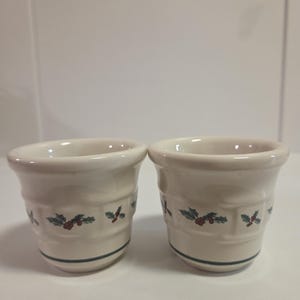Könnte beinhalten: Zwei cremefarbene Eierbecher aus Keramik. Jeder Becher hat ein grünes und rotes Stechpalmen-Beeren-Design in der Mitte und eine dunkelblaue Linie in der Nähe des Bodens. Die Becher haben einen leicht ausgestellten Rand.