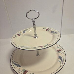 May include: Two-tiered serving stand with a silver handle and post. The off-white plates have a dark blue border and are decorated with floral patterns in red, blue, and green. Ideal for serving desserts or appetizers.
