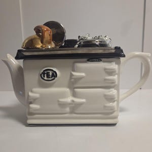 Könnte beinhalten: Eine weiße Keramik-Teekanne in Form eines Vintage-Küchenherds. Auf der Vorderseite befindet sich das Wort TEA in einem kreisförmigen Emblem. Die Oberseite enthält einen goldfarbenen Kessel und schwarze Akzente. Sie hat einen geschwungenen Griff rechts und einen Ausguss links.