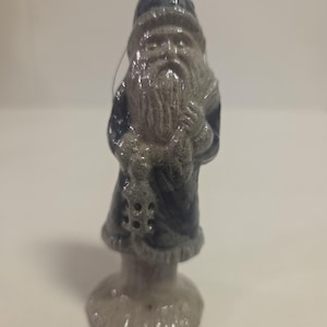 May include: A small, standing figurine of a bearded figure wearing a pointed hat and a long coat. The figure is rendered in a glossy, dark blue and grey speckled finish, holding a small lantern in one hand and a sack over the shoulder.