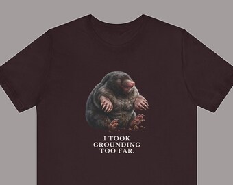 May include: A dark brown short-sleeve t-shirt featuring a graphic of a round, dark-furred mole with pink claws sitting on the ground. Below the mole, the white text reads: I TOOK GROUNDING TOO FAR. The shirt has a crew neckline and a standard fit.