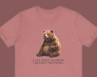 May include: A dusty rose short-sleeved t-shirt featuring a graphic of a brown bear sitting down. Below the bear, the text reads: I ATE NINE SALMON. I REGRET NOTHING. The shirt has a crew neckline and a standard fit, showcasing a simple design.