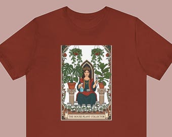 May include: A rust-colored t-shirt featuring a graphic illustration of a person wearing a crown, sitting on a throne between two large potted plants, holding a spray bottle and a moisture meter, with small jars of plants and the text THE HOUSE PLANT COLLECTOR.