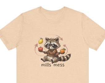 May include: A light tan short-sleeve t-shirt featuring a graphic of a raccoon wearing a brown striped jumper and glasses, juggling a yellow lemon, a red apple, and an orange. The text "mills mess" is printed in brown below the raccoon.