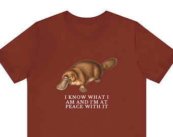 May include: A rust-colored short-sleeve t-shirt featuring a detailed illustration of a brown platypus. Below the animal, the text reads: I KNOW WHAT I AM AND I'M AT PEACE WITH IT. The shirt has a crew neckline and a standard fit, showcasing a graphic design.