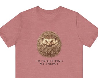 May include: A dusty rose short-sleeved t-shirt featuring a central graphic of a curled-up brown hedgehog. Below the hedgehog, the text reads "I'M PROTECTING MY ENERGY" in a dark, serif font. The shirt has a crew neckline and a heathered texture.