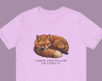 May include: A light purple short-sleeve t-shirt featuring a graphic of a curled-up orange fox. Below the fox, the text reads: I GREW THIS PILLOW. I'M USING IT. The shirt has a heathered texture and a standard crew neckline design.