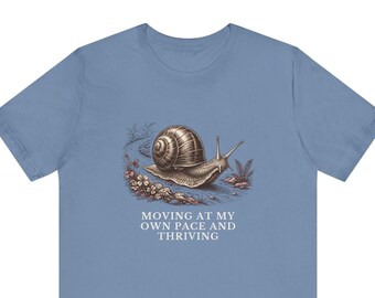 May include: A dusty blue t-shirt featuring a detailed brown illustration of a snail crawling on a path with small plants and rocks. Below the snail, the text reads: MOVING AT MY OWN PACE AND THRIVING in white capital letters.