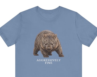 May include: A dusty blue short-sleeve t-shirt featuring a realistic illustration of a brown wombat centered on the chest. Below the animal, the text "AGGRESSIVELY FINE" is printed in a clean, white, sans-serif font against the blue fabric.