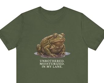 May include: An olive green t-shirt featuring a graphic of a brown toad sitting in a small puddle. Below the toad, the text reads: UNBOTHERED. MOISTURIZED. IN MY LANE. The toad has a textured, bumpy appearance and is depicted in a realistic style.