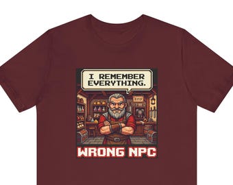 May include: A maroon short-sleeve t-shirt featuring a pixel art graphic of a bearded shopkeeper with arms crossed. A speech bubble above the character reads, "I REMEMBER EVERYTHING." Below the graphic, the text "WRONG NPC" is printed in red.