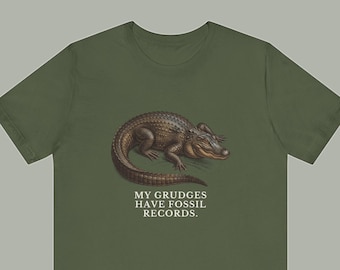 May include: An olive green short-sleeve t-shirt featuring a detailed illustration of a brown alligator in the center. Below the alligator, the text "MY GRUDGES HAVE FOSSIL RECORDS." is printed in a clean, white, capitalized serif font.